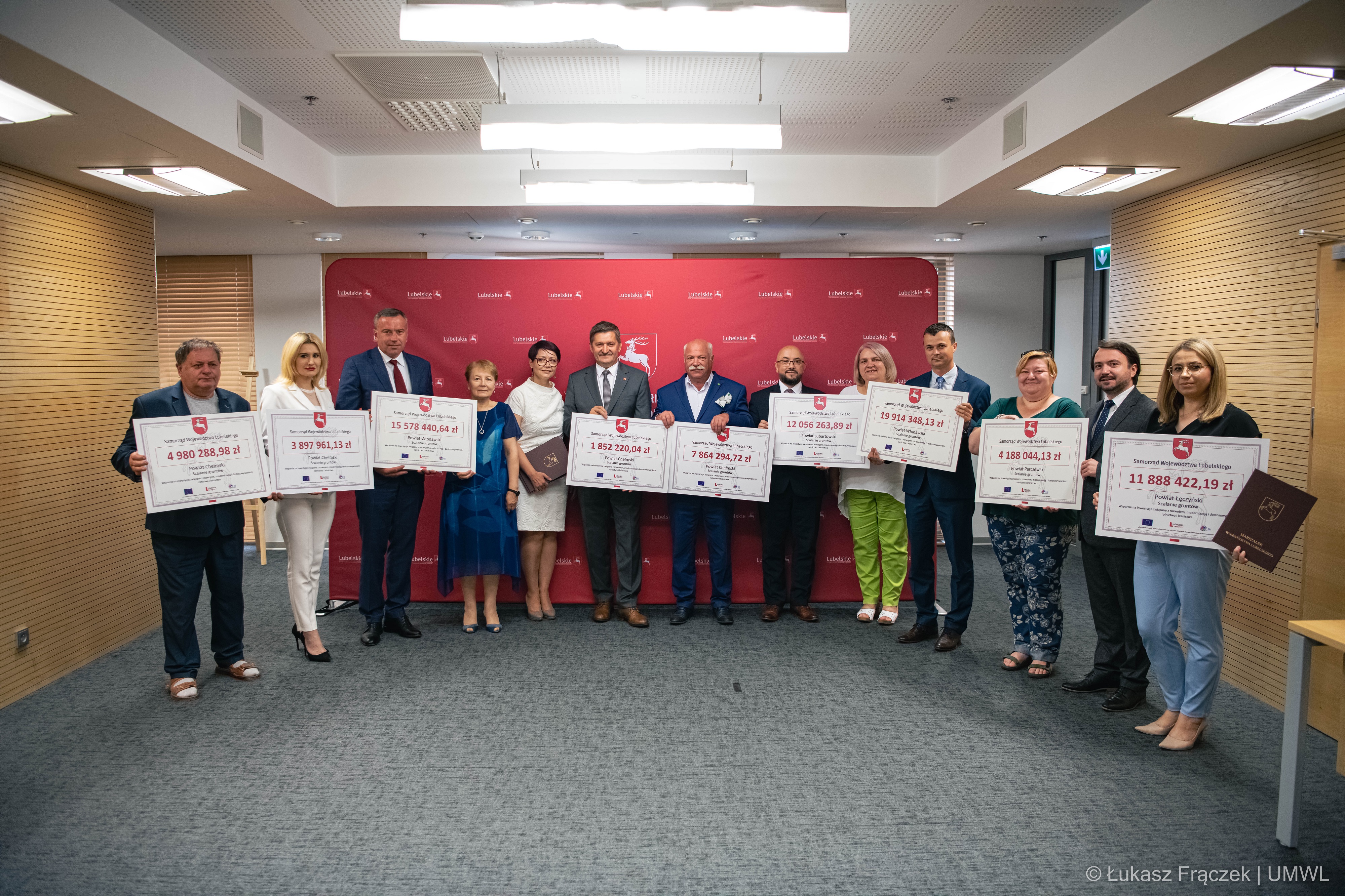 Widok na salę konferencyjną. Stoją w niej przedstawiciele wszystkich powiatów którzy trzymają w rękach tablice z wypisanymi kwotami uzyskanych dotacji. Pośrodku nich stoi Członek Zarządu Województwa Zdzisław Szwed