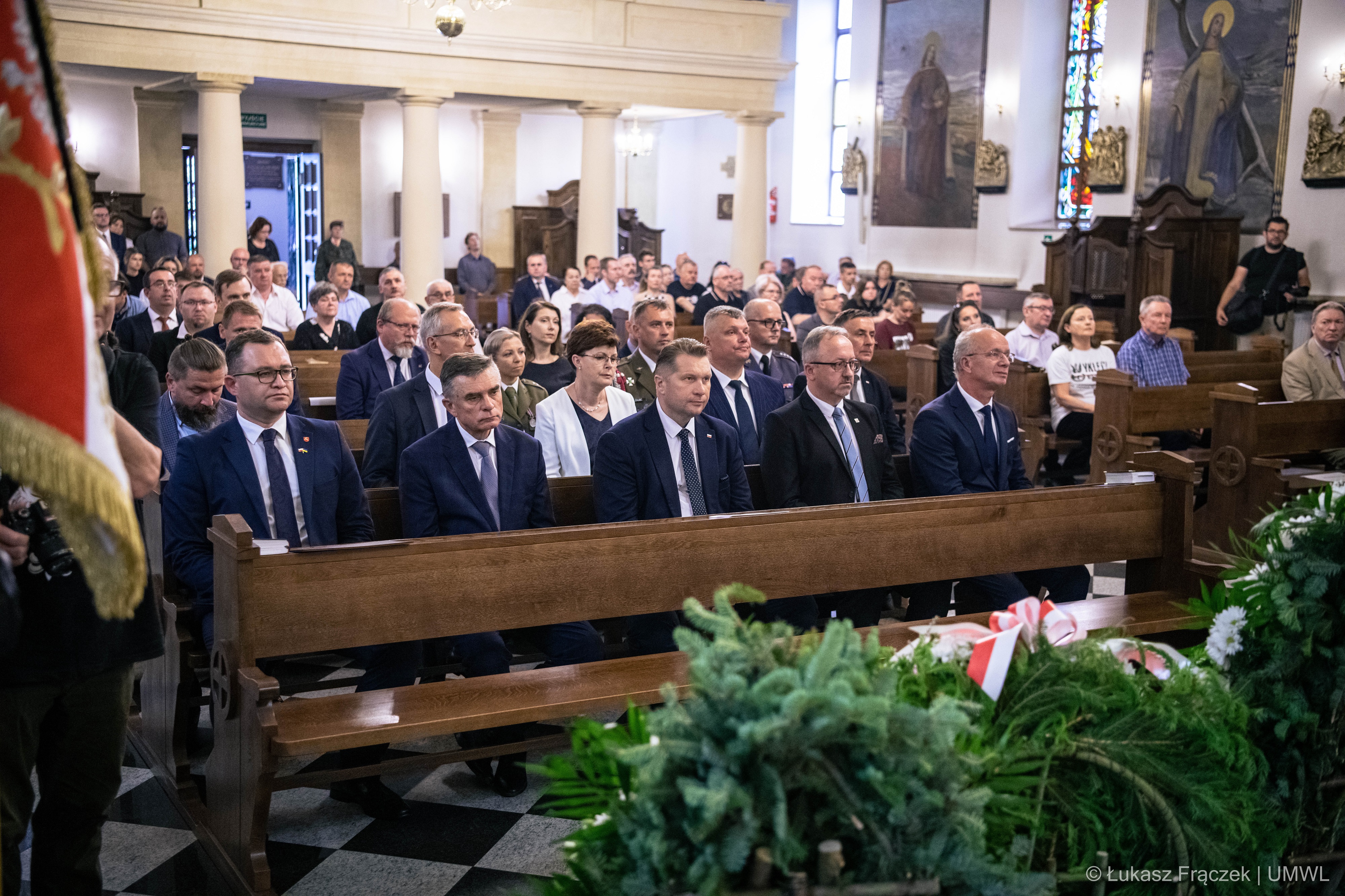 Widok na wnętrze kościoła i ludzi siedzących w ławkach. Na pierwszym planie widoczny fragment wiązanki pogrzebowej.