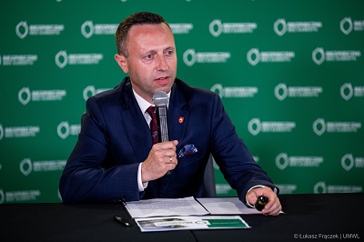 Mężczyzna w garniturze siedzi przy stole konferencyjnym, trzyma w ręce mikrofon i udziela wypowiedzi. Przed nim na stole leża notatki. W tle ścianka z napisem Bezpieczni bez azbestu