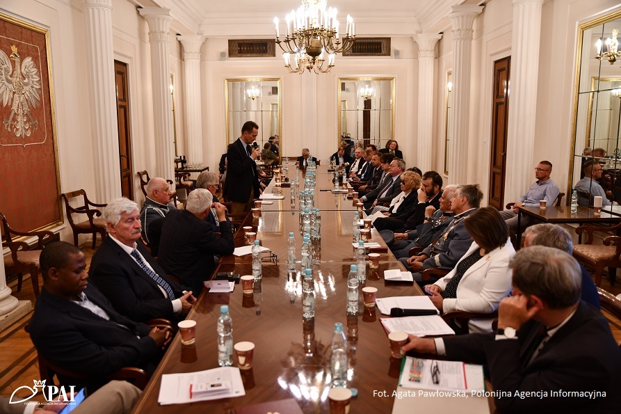Widok na długi stół konferencyjny, po jego obu stronach siedzą usadzeni goście, różnej płci i wieku, przed każdym z nich na stole stoi woda oraz leżą notatki