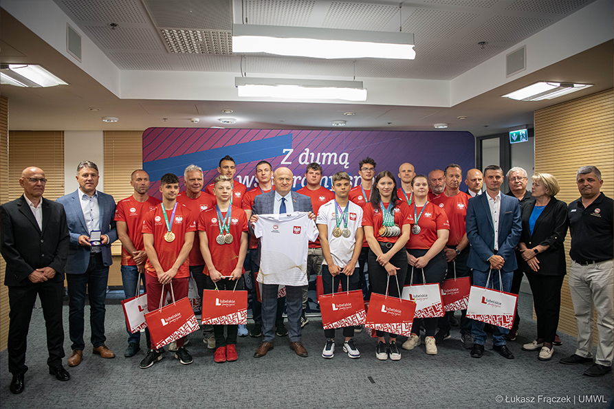 Wszyscy sportowcy, trenerzy uczestnicy spotkania oraz Marszałek Jarosław Stawiarski. Zdjęcie grupowe na tle fioletowo niebieskiej ścianki z napisem "Z dumą wspieramy sport". Marszałek Jarosław Stawiarski trzyma prezent w postaci koszulki.