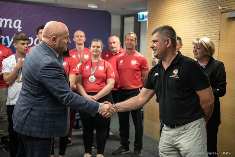 Marszałek Jarosław Stawiarski ściska dłoń trenera. W tle Sportowcy w czerwonych koszulkach