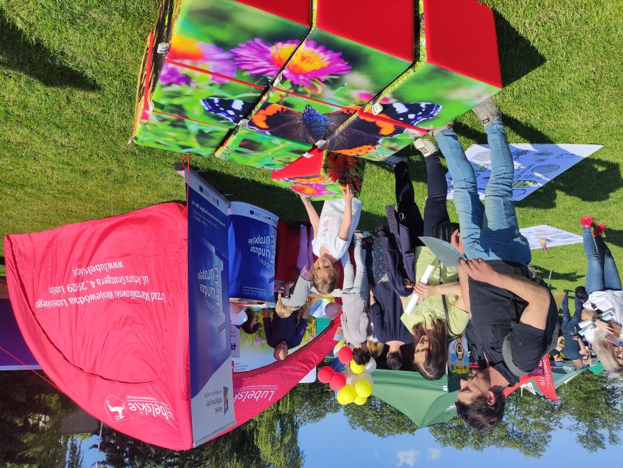 Układanie puzzli - dużych kolorowych kostek - przez małą dziewczynkę i jej rodziców. W tle namiot UMWL, balony oraz publiczność przechodząca między stoiskami