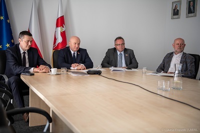 Na pierwszym planie stół przy którym siedzi czterech meżczyzn uczestniczących w wideokonferencji