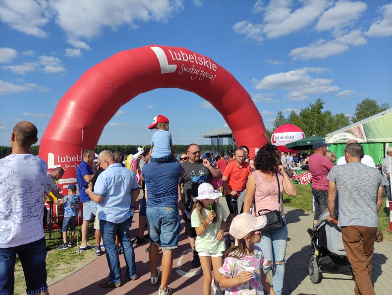 Brama promocyjna w kształcie półkola z napisem "Lubelskie - smakuj życie" pod którą przechodzi tłum ludzi. W górnej części zdjęcia błękitne niebo z obłokami.