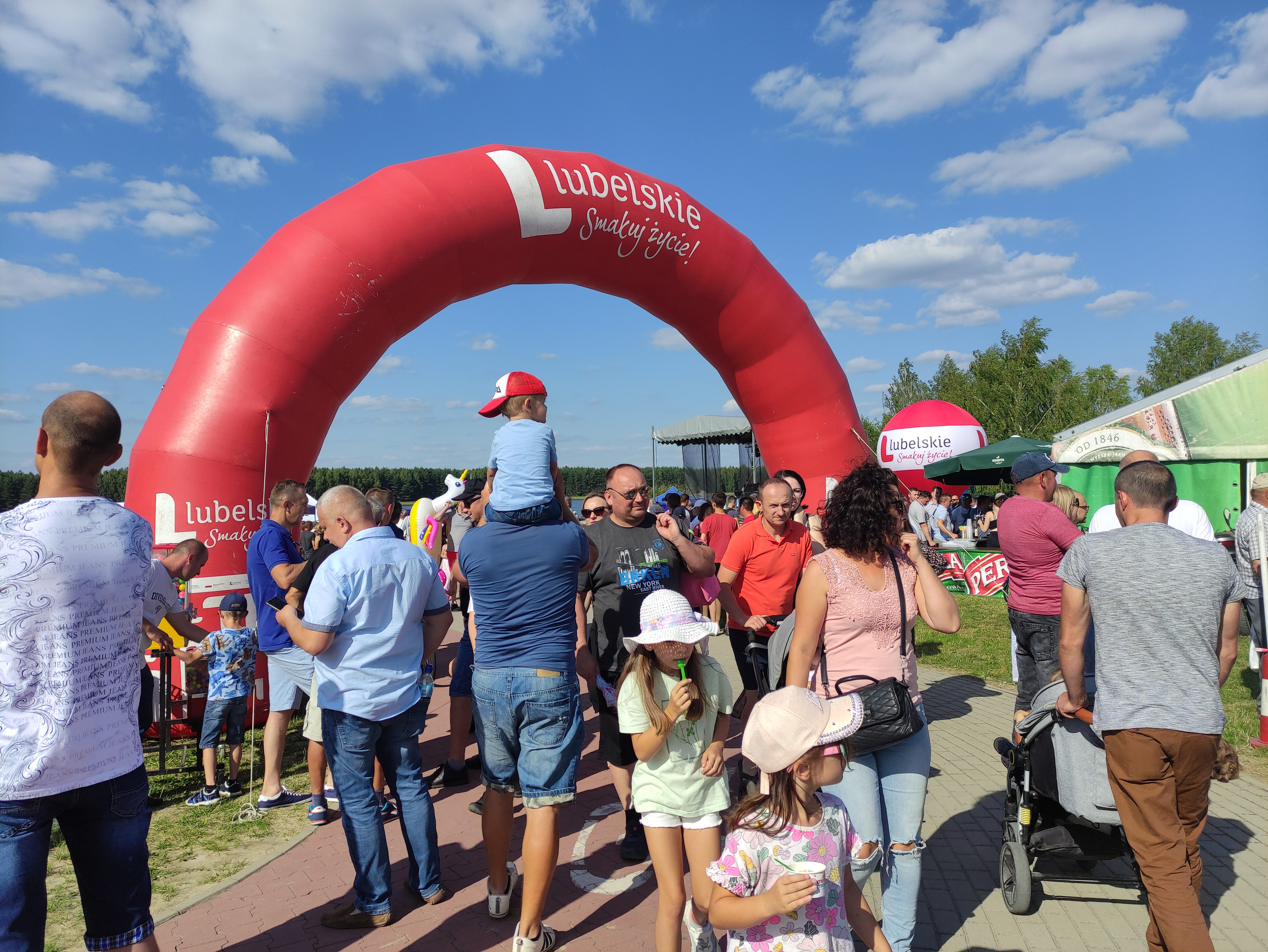 Brama promocyjna w kształcie półkola z napisem "Lubelskie - smakuj życie" pod którą przechodzi tłum ludzi. W górnej części zdjęcia błękitne niebo z obłokami.