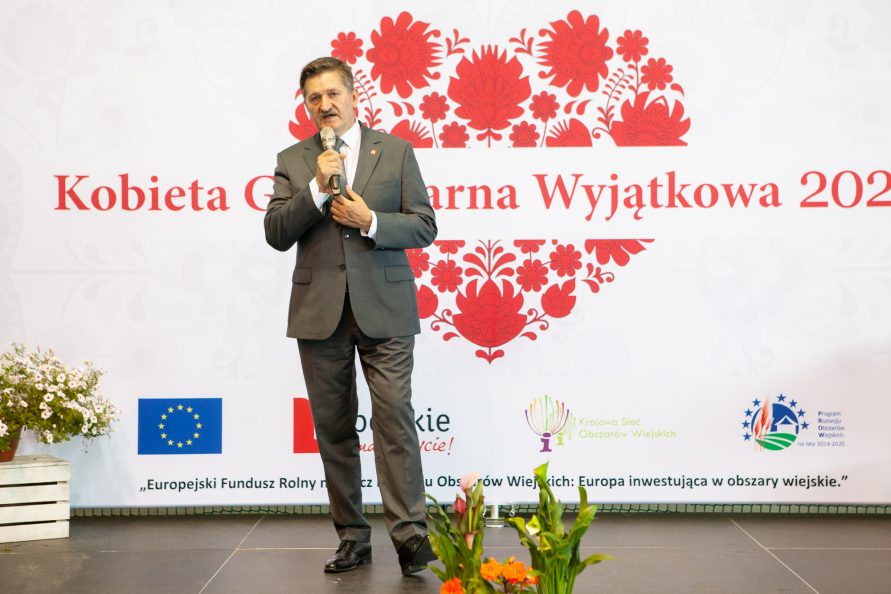 Członek Zarządu Województwa Lubelskiego Zdzisław Szwed przemawiający podczas półfinału konkursu Kobieta Gospodarna Wyjątkowa 2022 w Chełmie. W tle czerwone serce wypełnione wzorami łowickimi. Przez jego środek na białym tle napis Kobieta Gospodarna Wyjątkowa. Poniżej logotypy: Unii Europejskiej, Lubelskie Smakuj Życie, KSOW oraz PROW 2014-2020. Pod logotypami napis: „Europejski Fundusz Rolny na Rzecz Rozwoju Obszarów Wiejskich: Europa inwestująca w obszary wiejskie”.