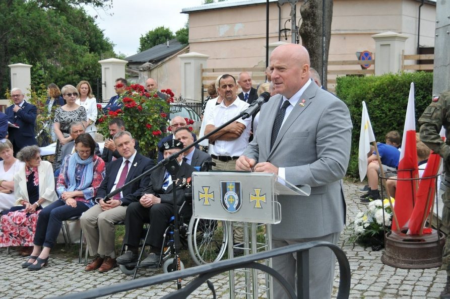 Mężyczna w garniturze stoi na placu pod kościołem i wygłasza przemówienie do zebranych tam osób. W tle widać siedzącego na krześle kombatanta