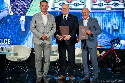 Trzech mężczyzn stoi w garniturach an scenie, dwóch z nich trzyma teczki. Za nimi ekran z trasą wyścigu rowerowego.