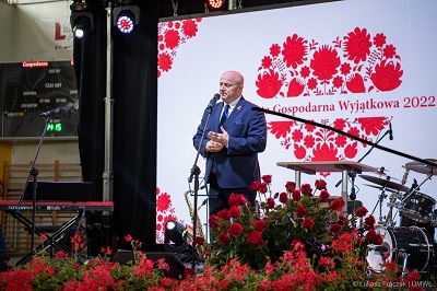 Marszałek Województwa Lubelskiego stoi na scenie przy mikrofonie za nim na ekranie wyświetla się logo wydarzenia Kobieta Gospodarna Wyjątkowa 2022