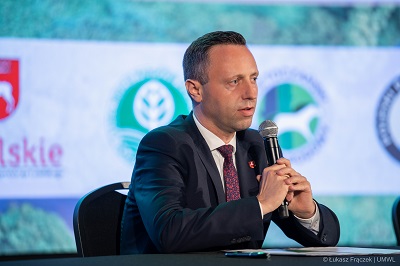 Mężczyzna siedzi przy stole trzyma mikrofon w obu rekach i udziela wypowiedzi, za nim duzy ekran