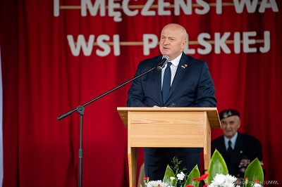 Marszałek WL stoi przy mikrofonie na scenia, za nim w tle widać siedzącego kombatanta i