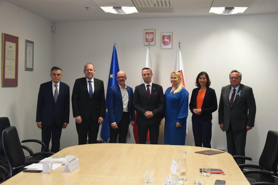 Siedem osób stoi obok siebie na tle trzech flag i białej ściany