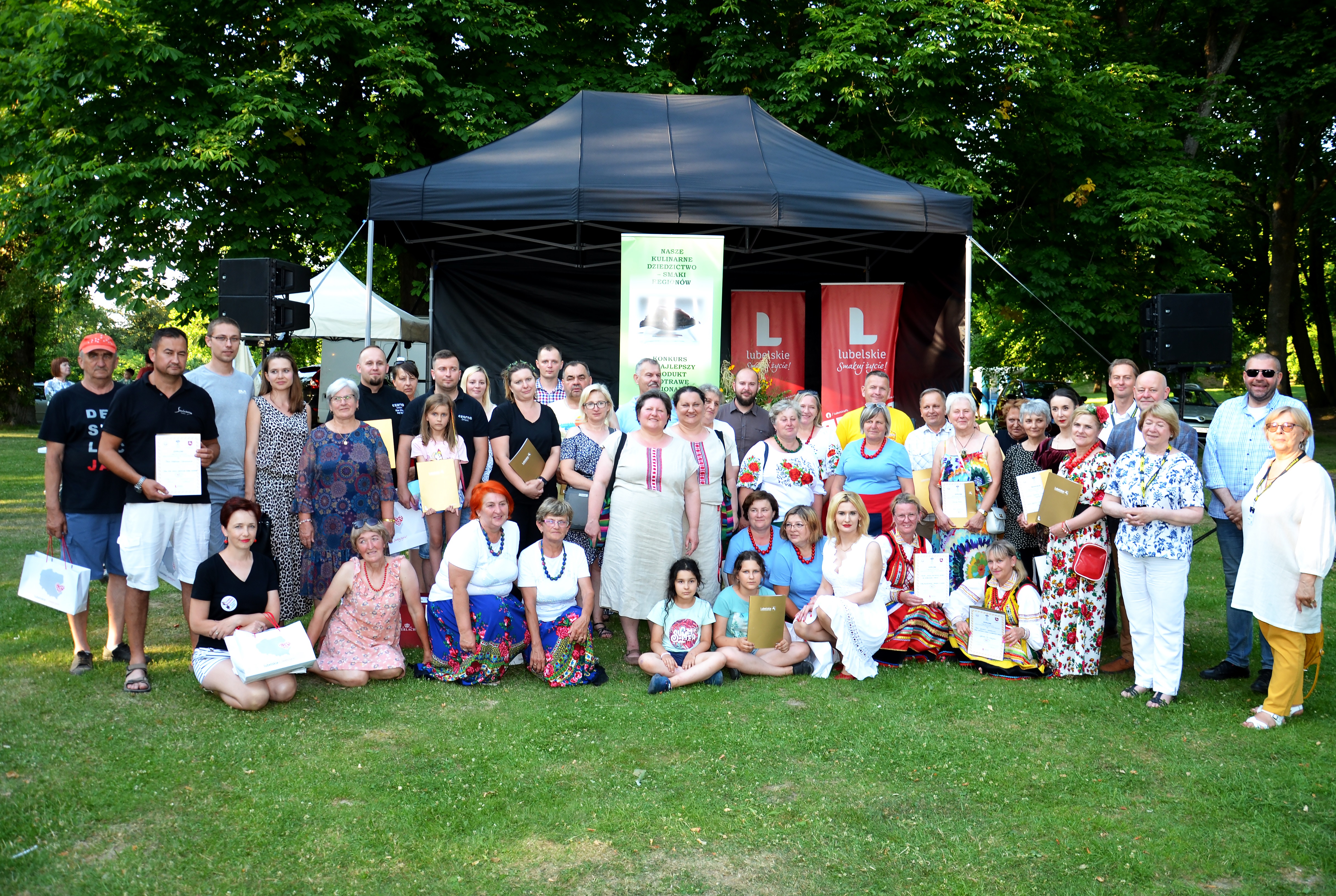 Grupowe zdjecie uczestników kinkursu kulinarnego,. Duża grupa osób w różnym wieku i płci, stoją na dworze podczas letniej pogody. Za nimi w tle widać rozstawiony namiot promocyjny z banerami reklamowymi