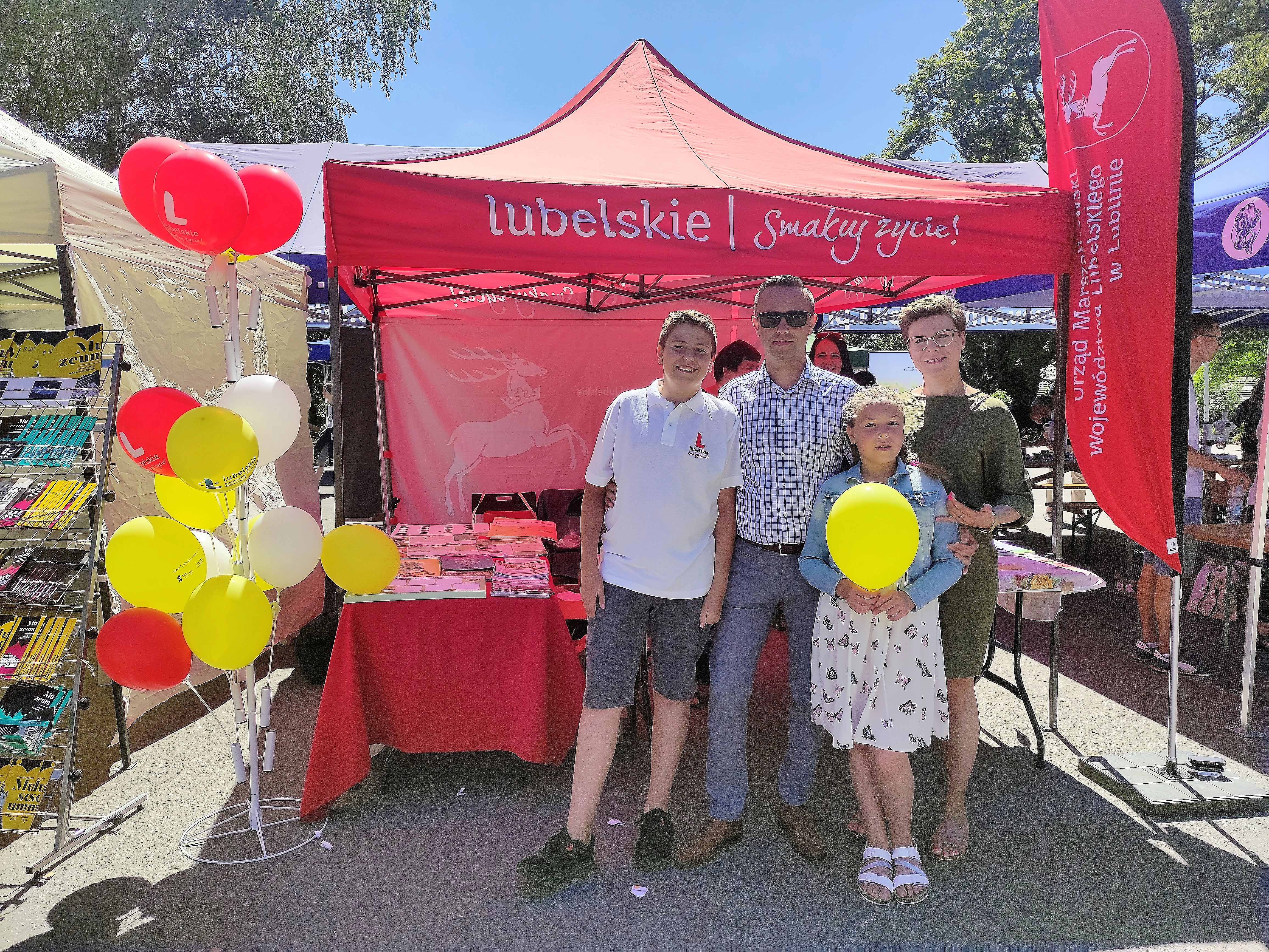 Rodzina (mama, tata i dwójka dzieci) przed namiotem wystawienniczym. Dziewczynka trzyma balon. Stojak z balonami widoczny po lewej stronie.