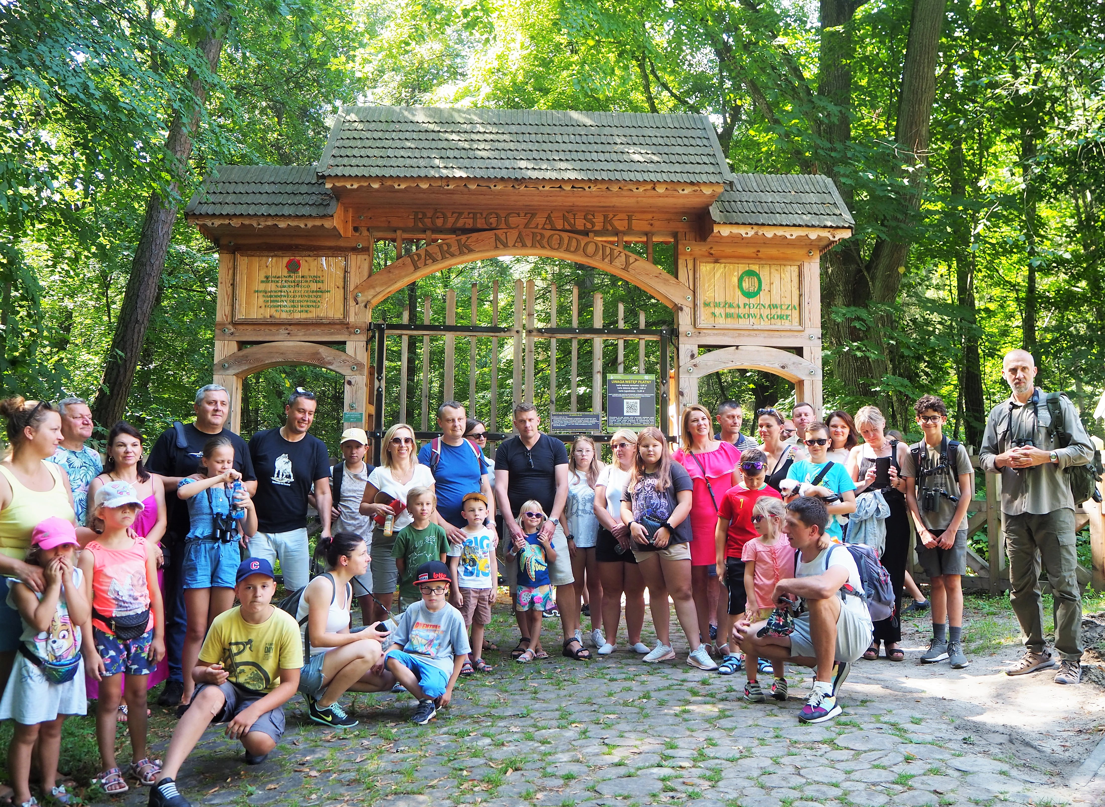 Grupa kilkudziesięciu osób w różnym wieku w strojach sportowych stoi i kuca pod wysoką drewnianą bramą z napisem: "Roztoczański Park Narodowy. Ścieżka poznawcza na Bukową Górę". W tle za bramą wysokie drzewa oświetlone promieniami słońca.
