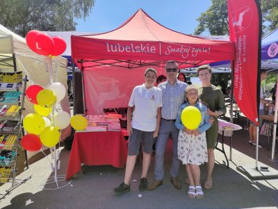 Rodzina (mama, tata i dwójka dzieci) przed namiotem wystawienniczym. Dziewczynka trzyma balon. Stojak z balonami widoczny po lewej stronie.