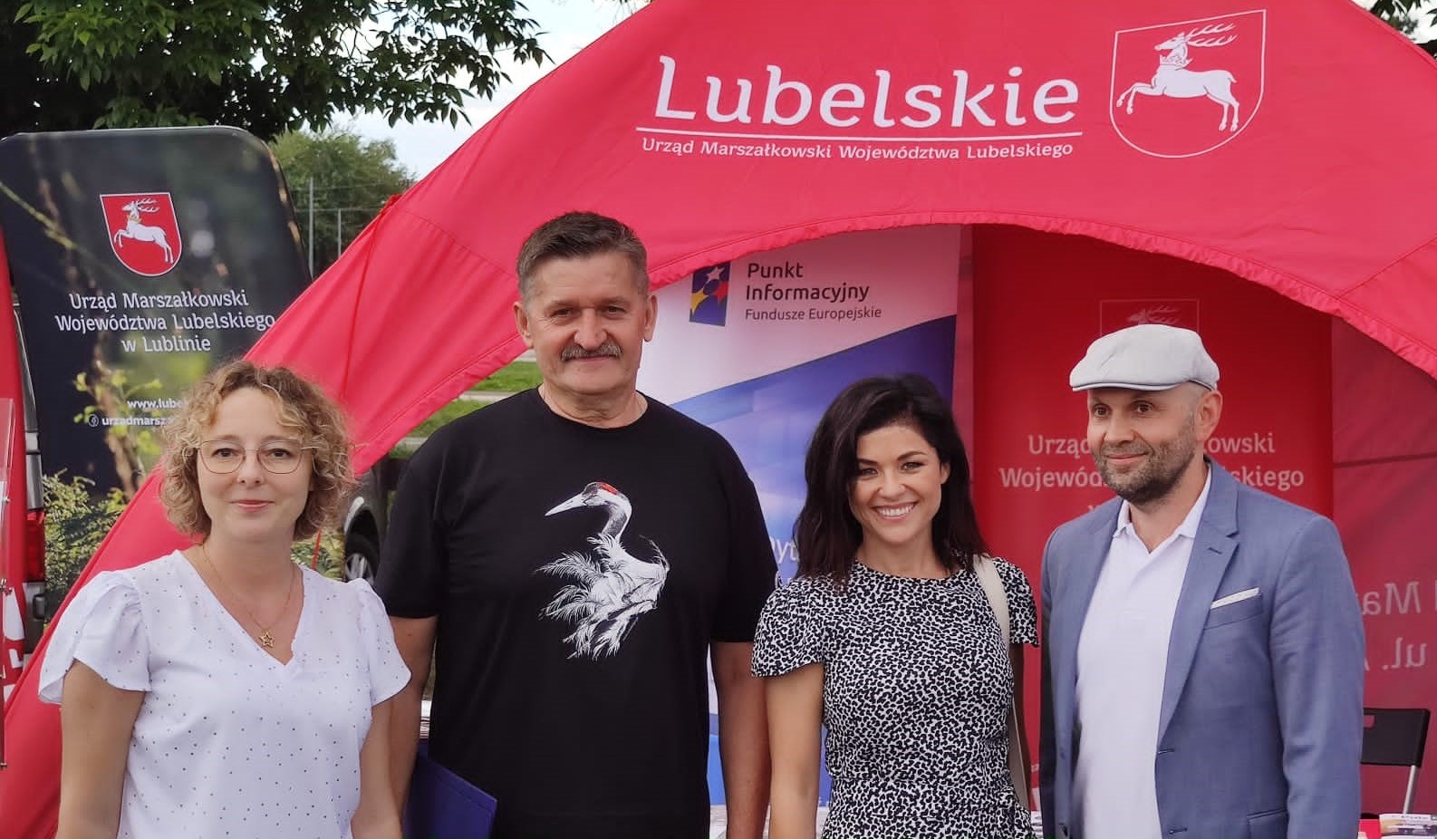 Osoby na tle stoiska Urzędu Marszałkowskiego. Cztery uśmiechnięte osoby, w tym dwie kobiety i dwóch mężczyzn, stoją przed namiotem z napisem "Lubelskie. Urząd Marszałkowski Województwa Lubelskiego". W tle po lewej widoczny fragment zadrzewień.