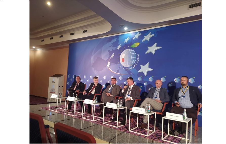 Kilku mężczyzn siedzi na krzesłach na scenie i biorą udział w trwającej debacie