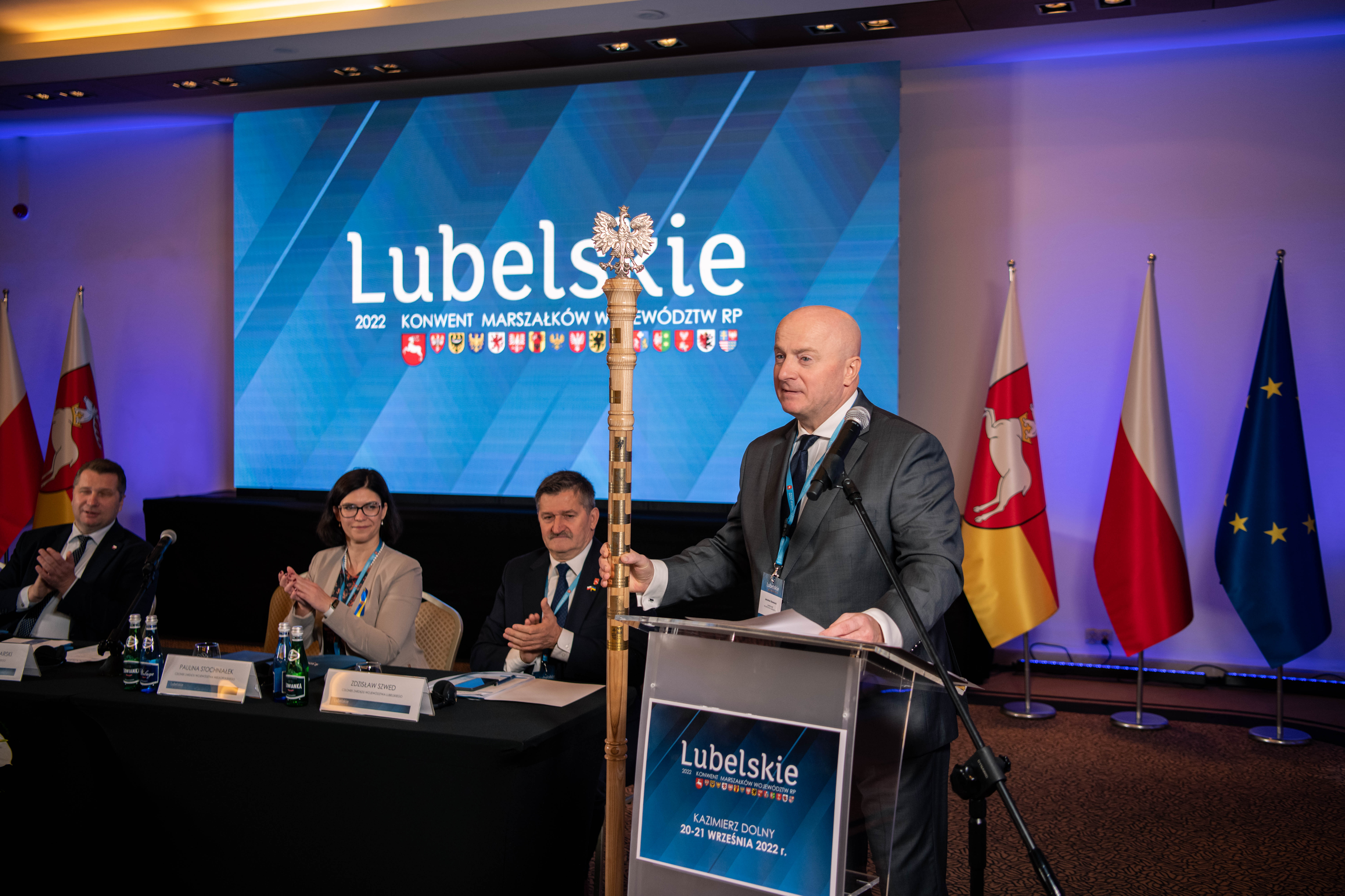 Marszałek Jarosław Stawiarski trzyma w ręku laskę marszałkowską stojąc przy mównicy. W tle niebieski ekran z napisem "Lubelskie", trzy flagi oraz trzy osoby siedzące przy stole