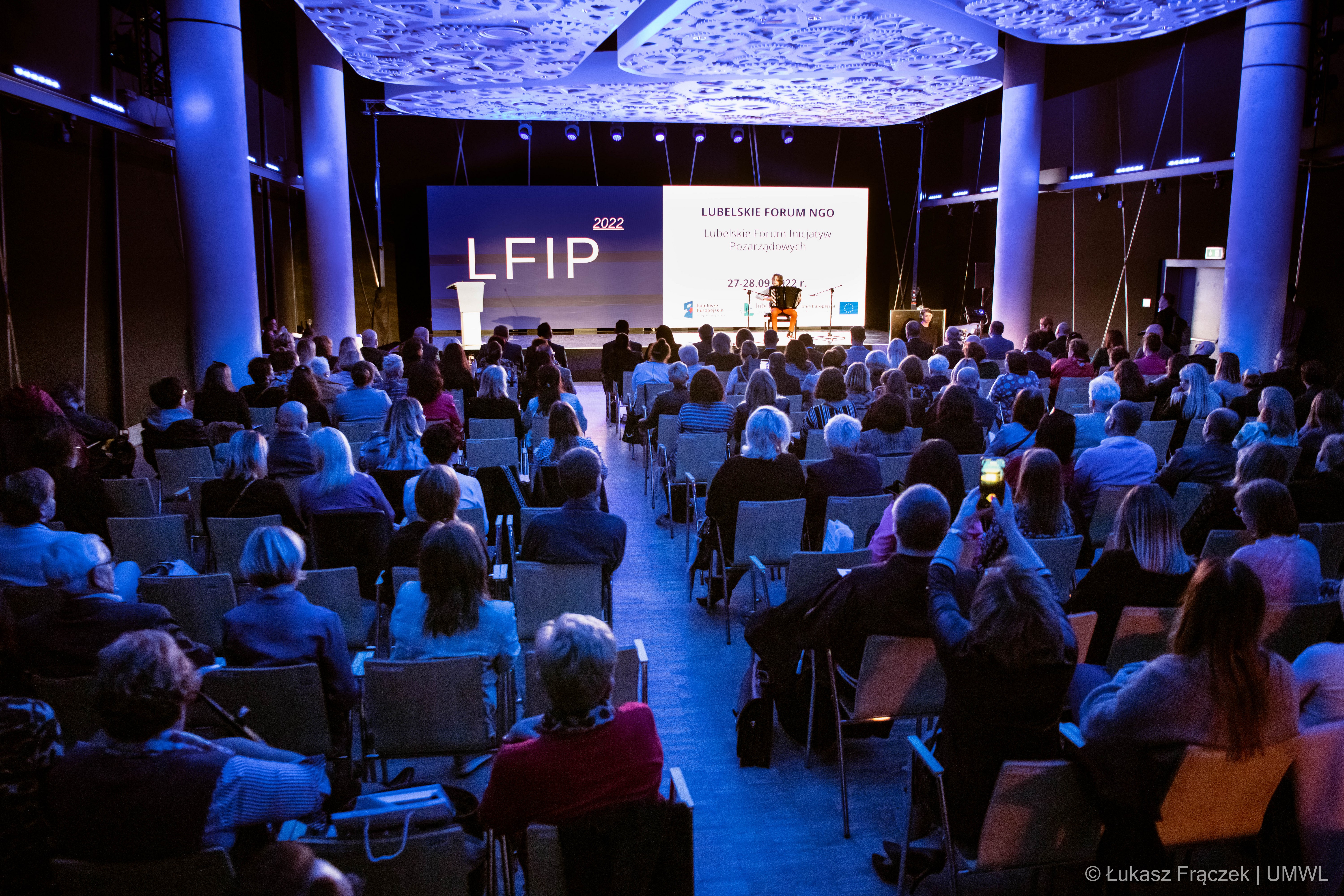 Na pierwszym planie publiczność zwrócona w stronę sceny. Dalej na scenie chłopiec grający na akordeonie na dla ekranu z napisem "LFIP 2022. Lubelskie forum NGO"