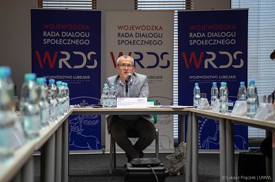 Widok na Przewodniczącego Zespołu, który siędzi przy stole i udziela wypowiedzi do mikrofonu. Po jego dwóch stronach stoją stoły w ustawionymi butelkami wody.