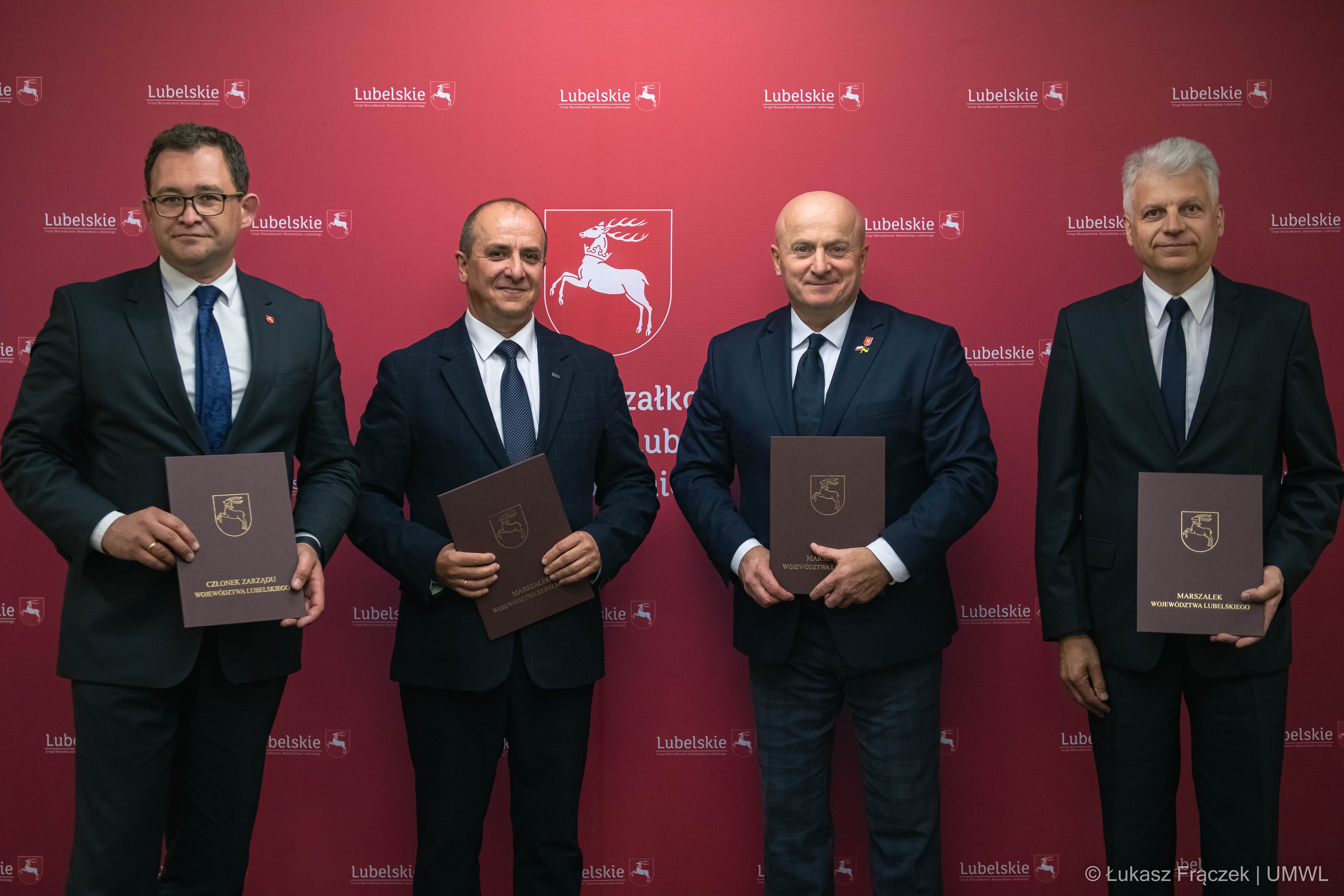 Czterech mężczyzn w tym marszałek WL stoją w sali konferencyjnej i trzymają w rękach teczki z podpisanymi umowami, za nimi ścianka UMWL