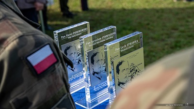 trzy szklane tabliczki pamiątkowe ustawione na stole na dworze,na pierwszym planie widoczny kawałek munduru stojącego żołnierza