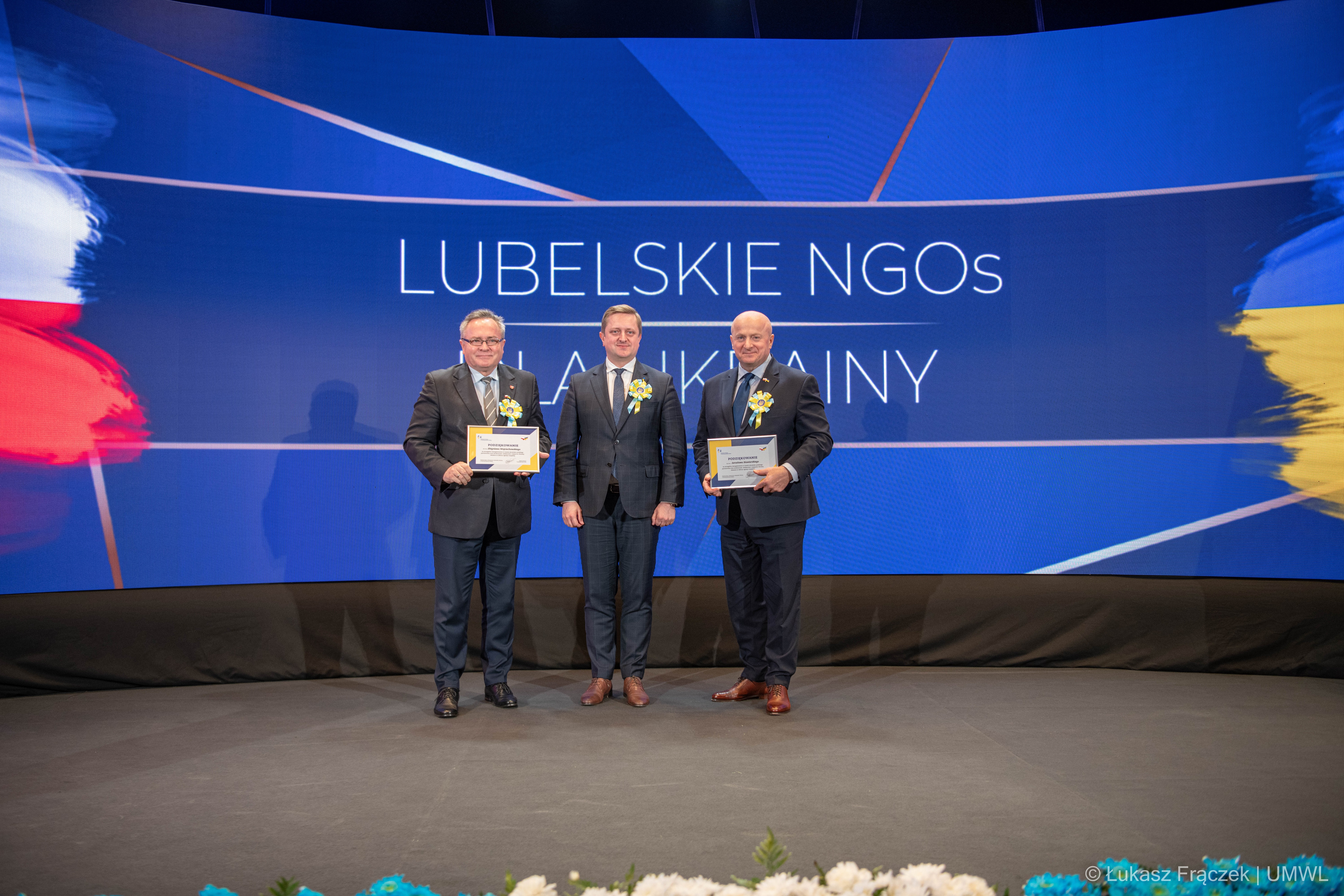 Marszałek WL, wicemarszałek WL i ambasador Ukrainy pozują wspólnie do zdjęcia na tle ekranu wyświetlającego napis Lubelskie NGOs dla Ukrainy