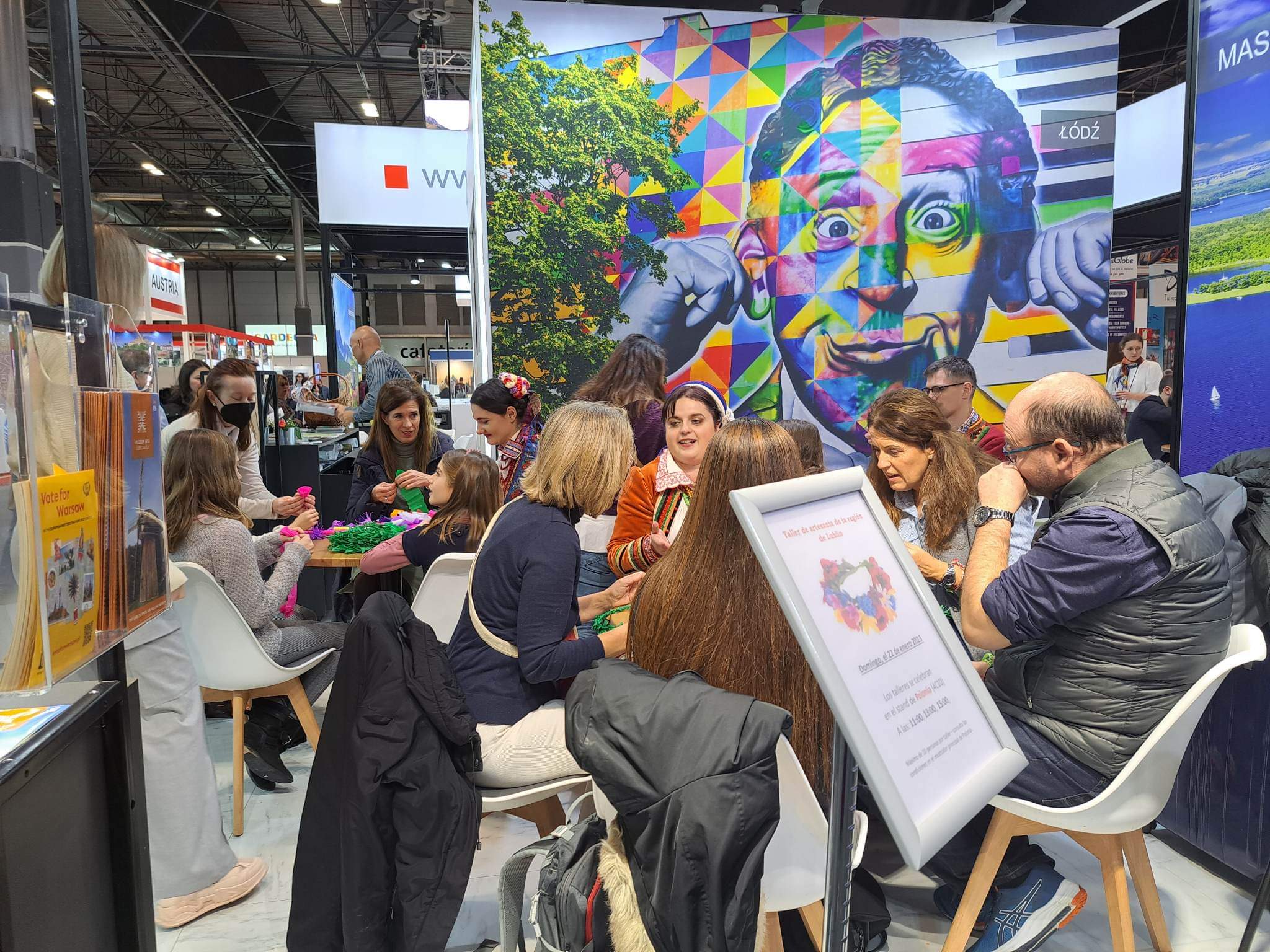 Sala w hali wypełniona ludźmi siedzącymi przy stolikach i wykonujących pod instruktażem ozdoby ludowe