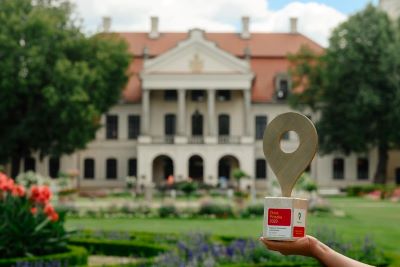 w tle pałac w Kozłówce od frontu, na pierwszym planie złota pinezka trzymana na dłoni