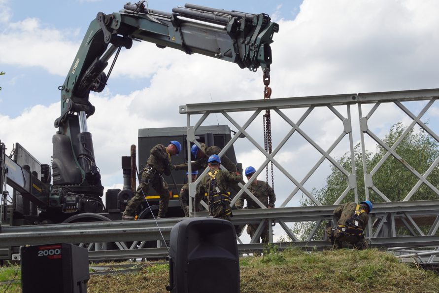 Widok na metalową konstrukcje mostu i żołnierzy, który sa w trakcie pracy nad stawianiem jej