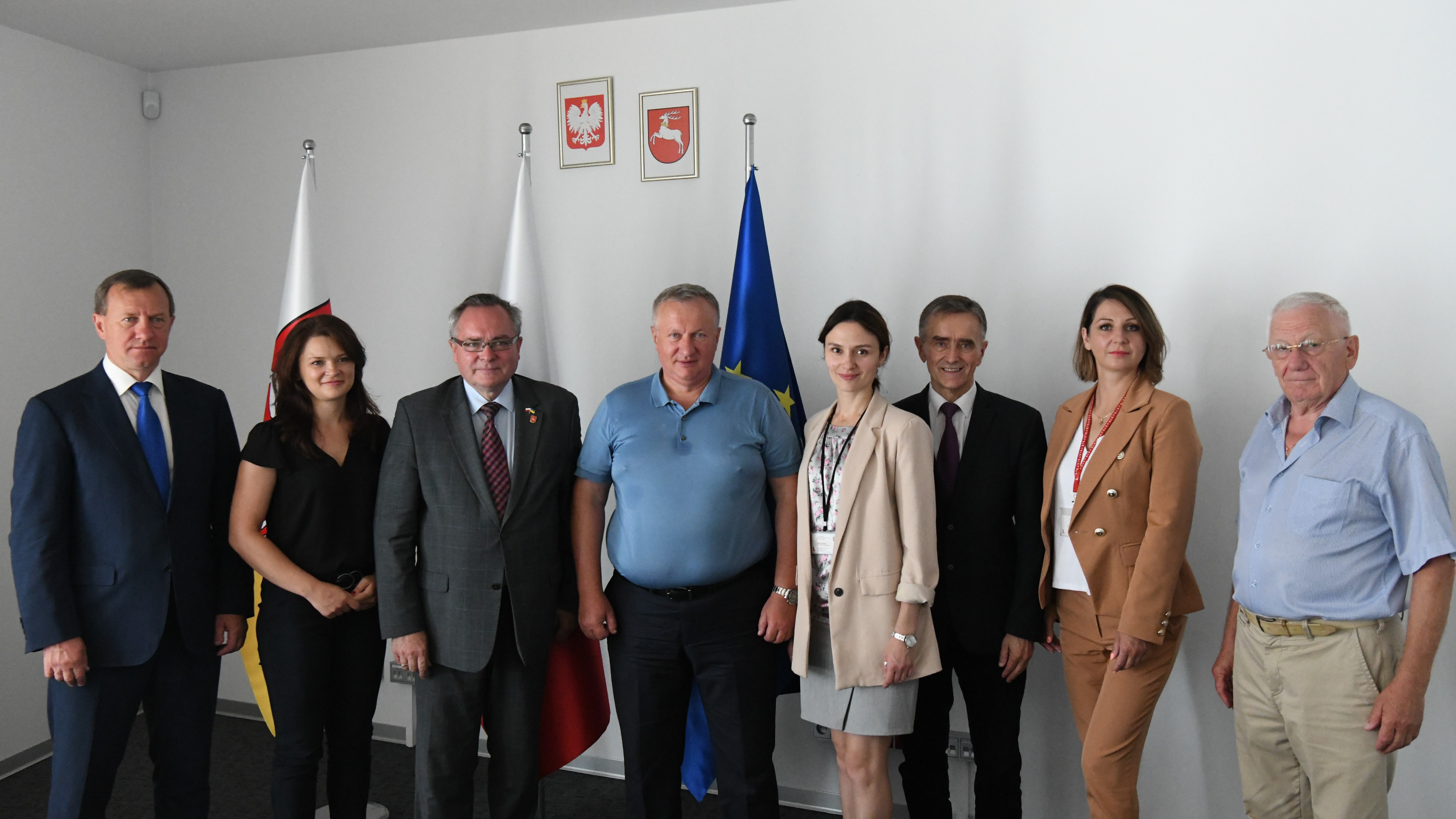 Wszyscy uczestnicy spotkania pozują do wspólnego zdjęcia. To około dziesięciu osób w większości mężczyzn, którzy stoją w zamkniętym pomieszczeniu. Za nimi znajdują się ustawione flagi