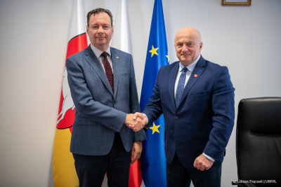 Marszałek Jarosław Stawiarski podaje rękę ambasadorowi Izraela. Za nimi znajdują się stojaki z flagą województwa, Polski i UE