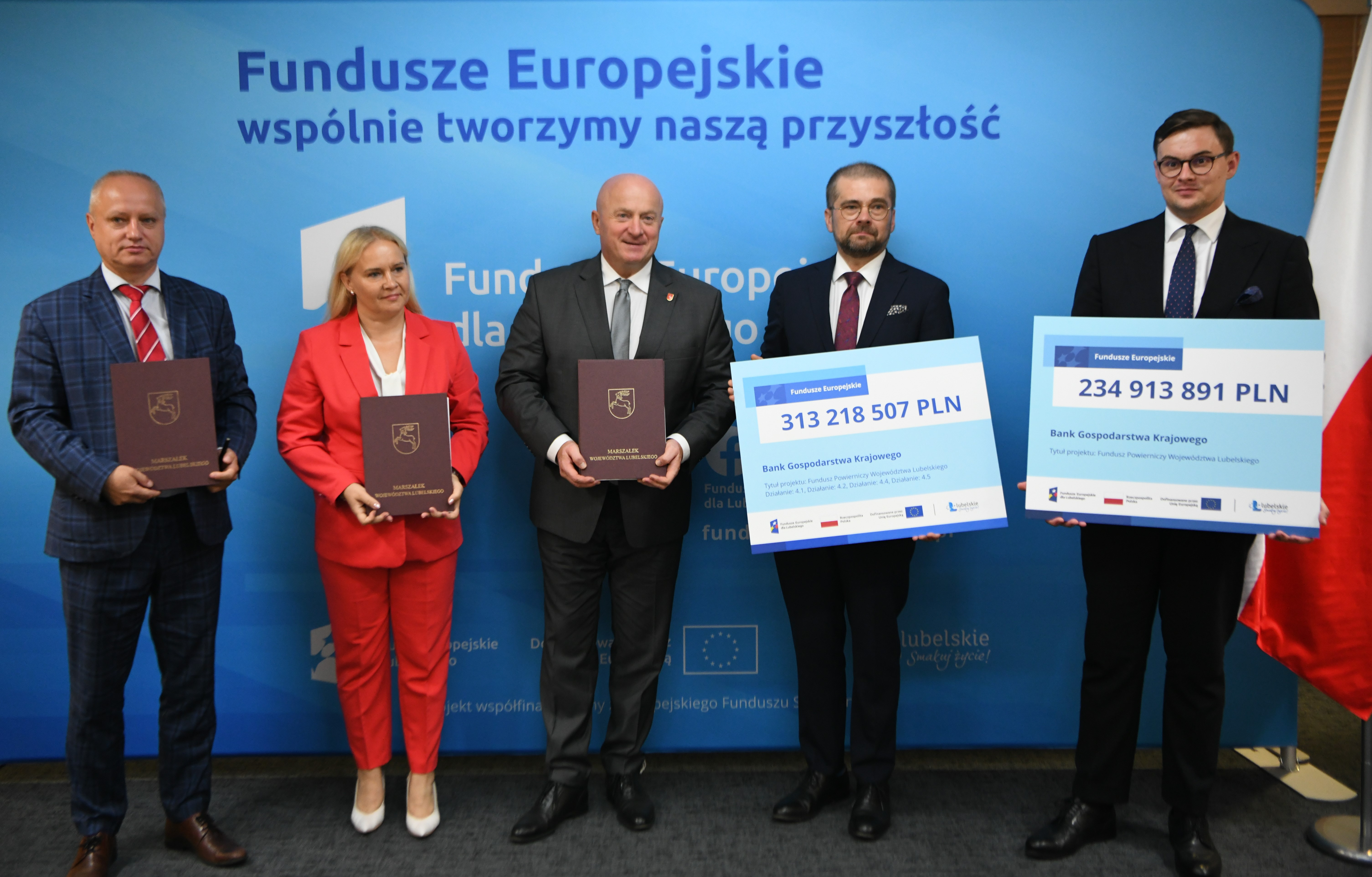 Uczestnicy spotkania czterech mężczyzn i jedna kobieta, wśród nich marszałek, stoją i pozują do zdjęcia trzymając w rękach symboliczne tablice z informacją o kwocie dofinansowania