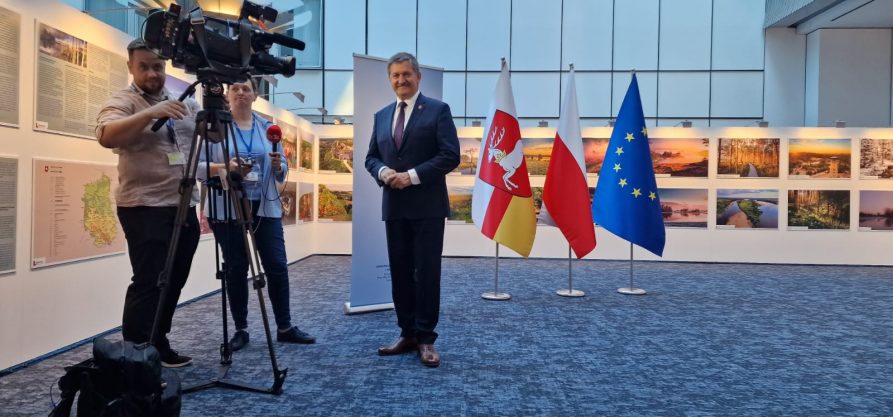 Członek Zarządu Zdzisław Szwed stoi na korytarzu wewnątrz nowoczesnego budynku, za nim stoją flagi i wystawa zdjęć