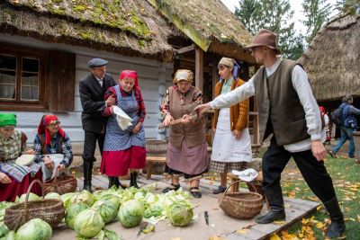 grupa kobiet i mężczyzn w ludowych strojach na tle chałupy Scena przygotowania kapusty do kiszenia