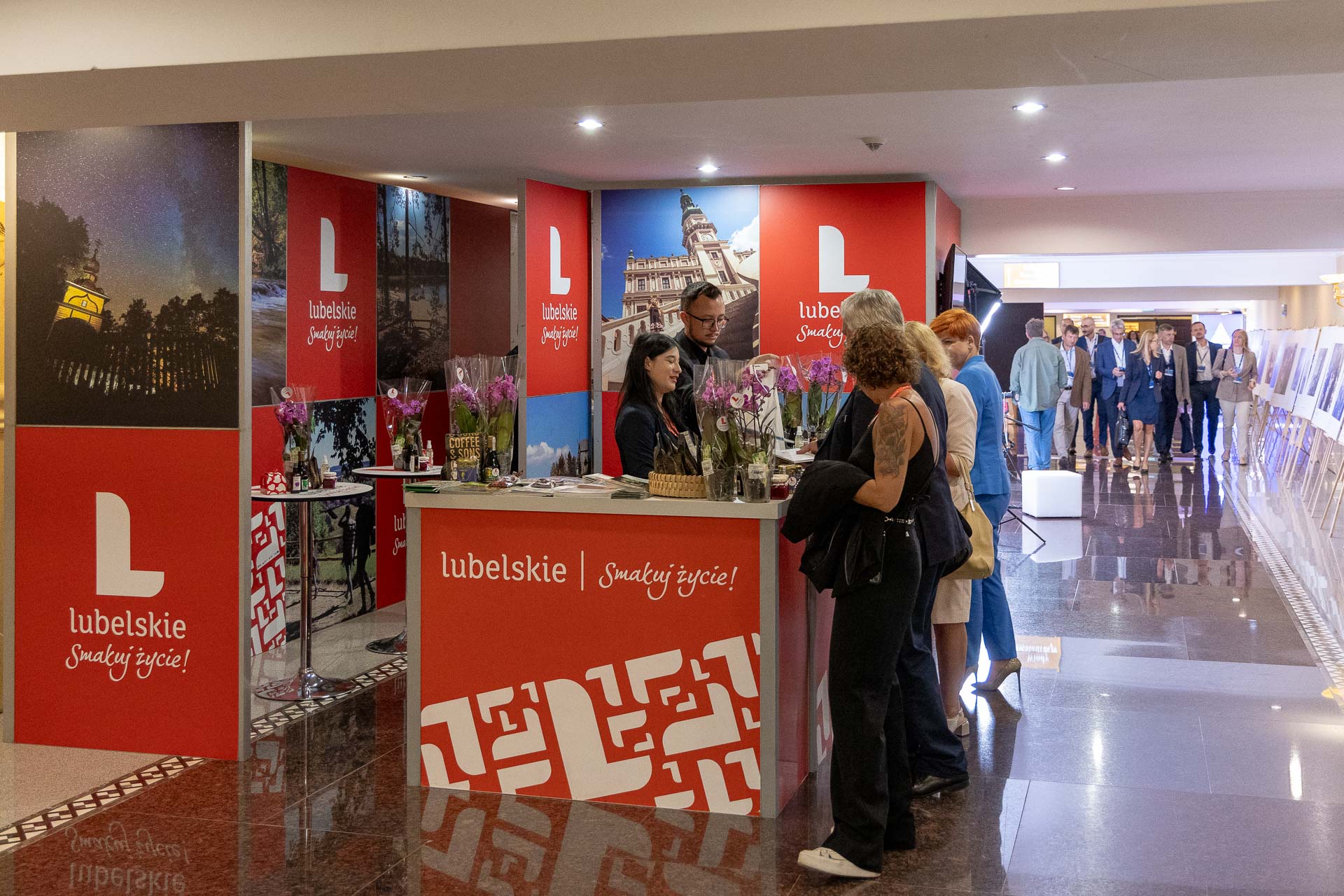 Wnętrze korytarza, zbliżenie na ustawione stoisko promocyjne przy którym stoją trzy osoby obsługujące i kilka zainteresowanych