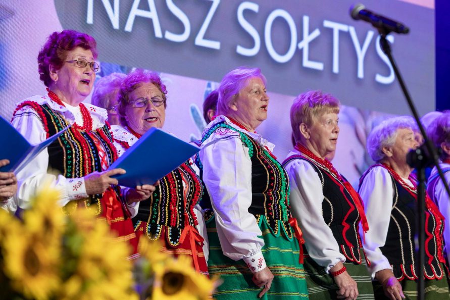 Panie w strojach ludowych stoją na scenie w trakcie swojego występu śpiewaczego