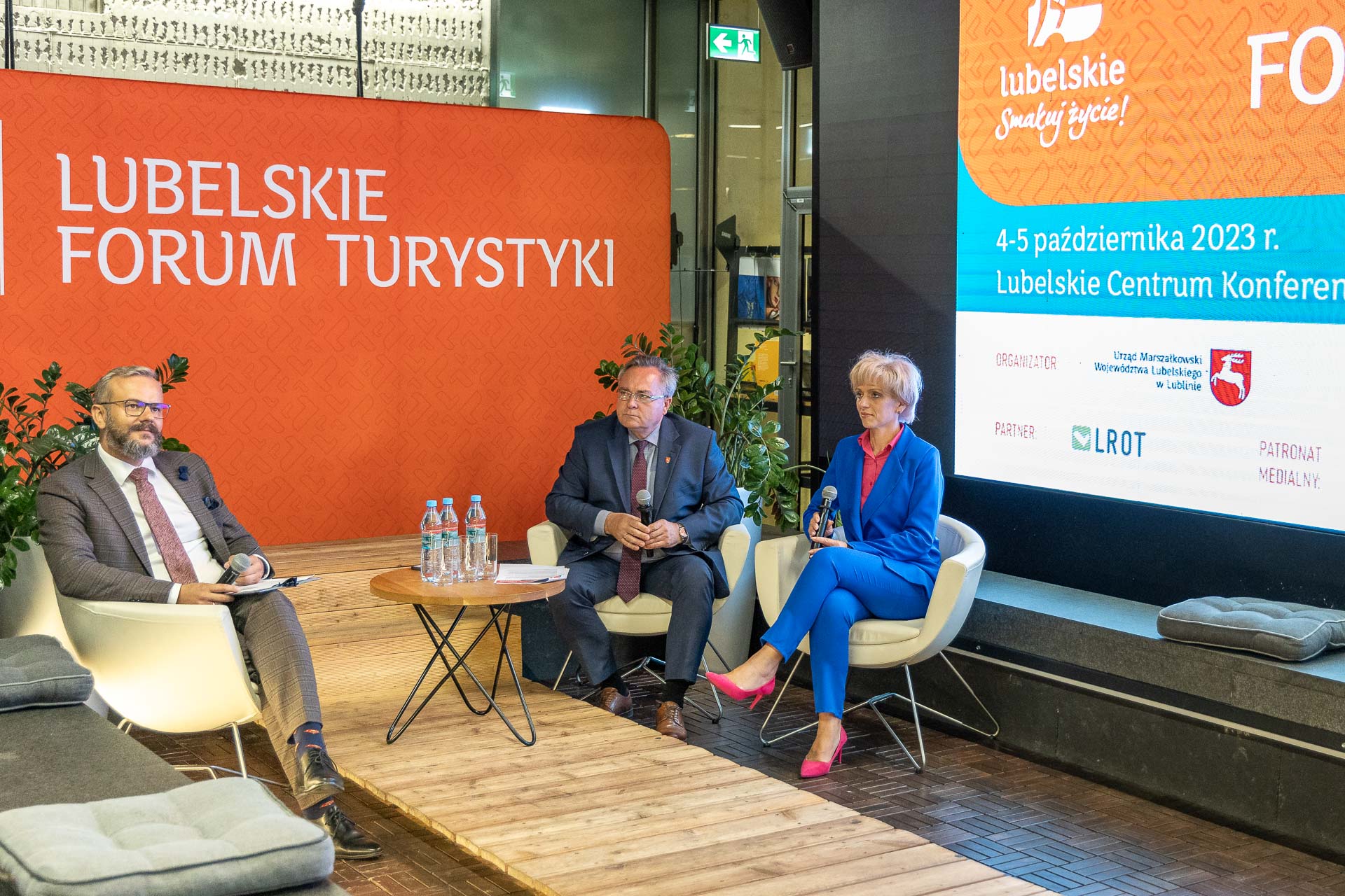 Troje uczestników briefingu prasowego . Wszyscy siedzą w fotelach, za nimi znajduje się ścianka z napisem Lubelskie Forum Turystyki