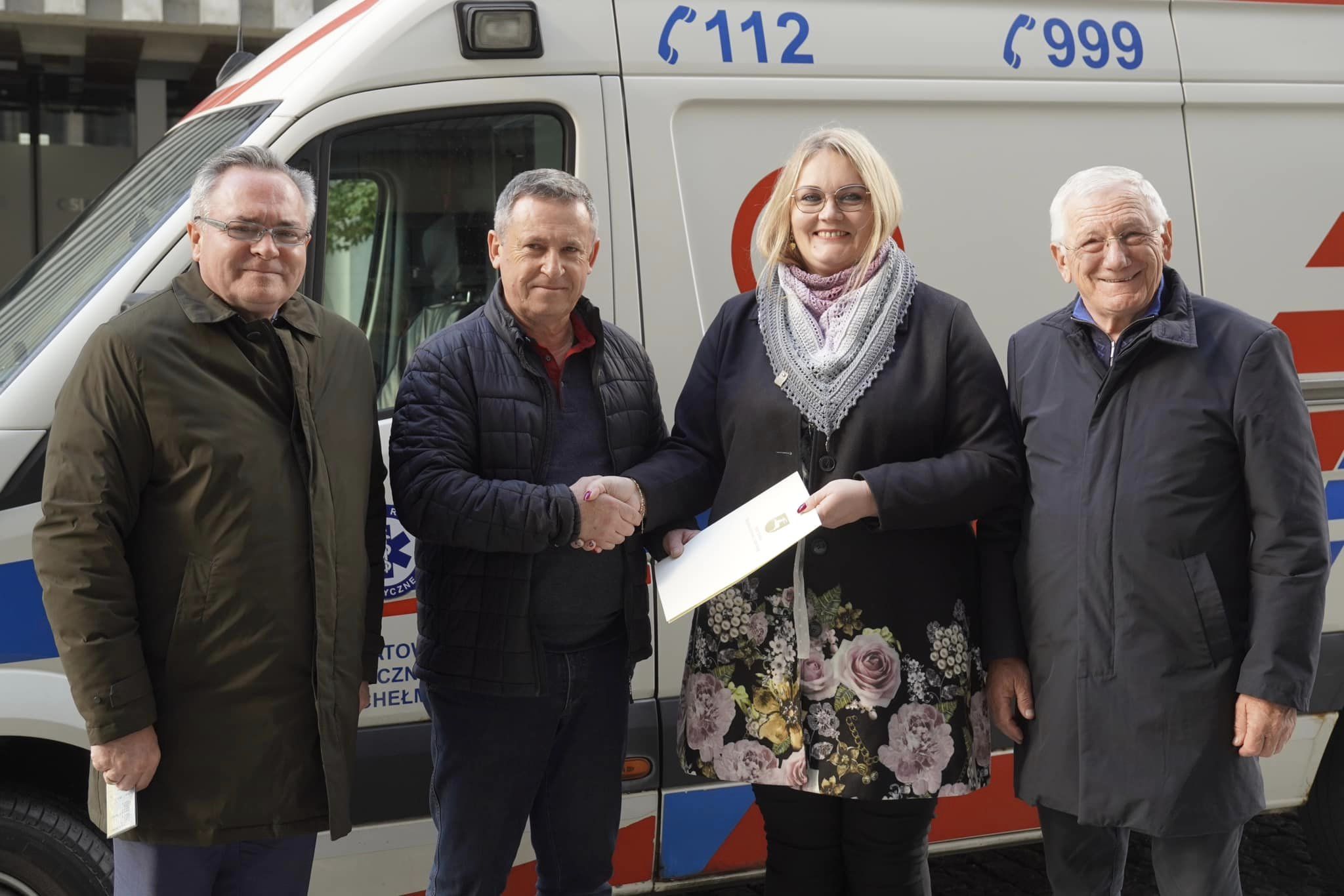 Cztery osoby na zdjęciu stoją na zewnątrz. Za nimi znajduje się ambulans. Jedna z osób, kobieta, podaje rękę stojącemu obok mężczyźnie