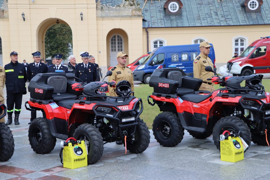 Quady ustawione na zewnątrz, przed budynkiem, przy każdym quadzie stoi strażak w mundurze