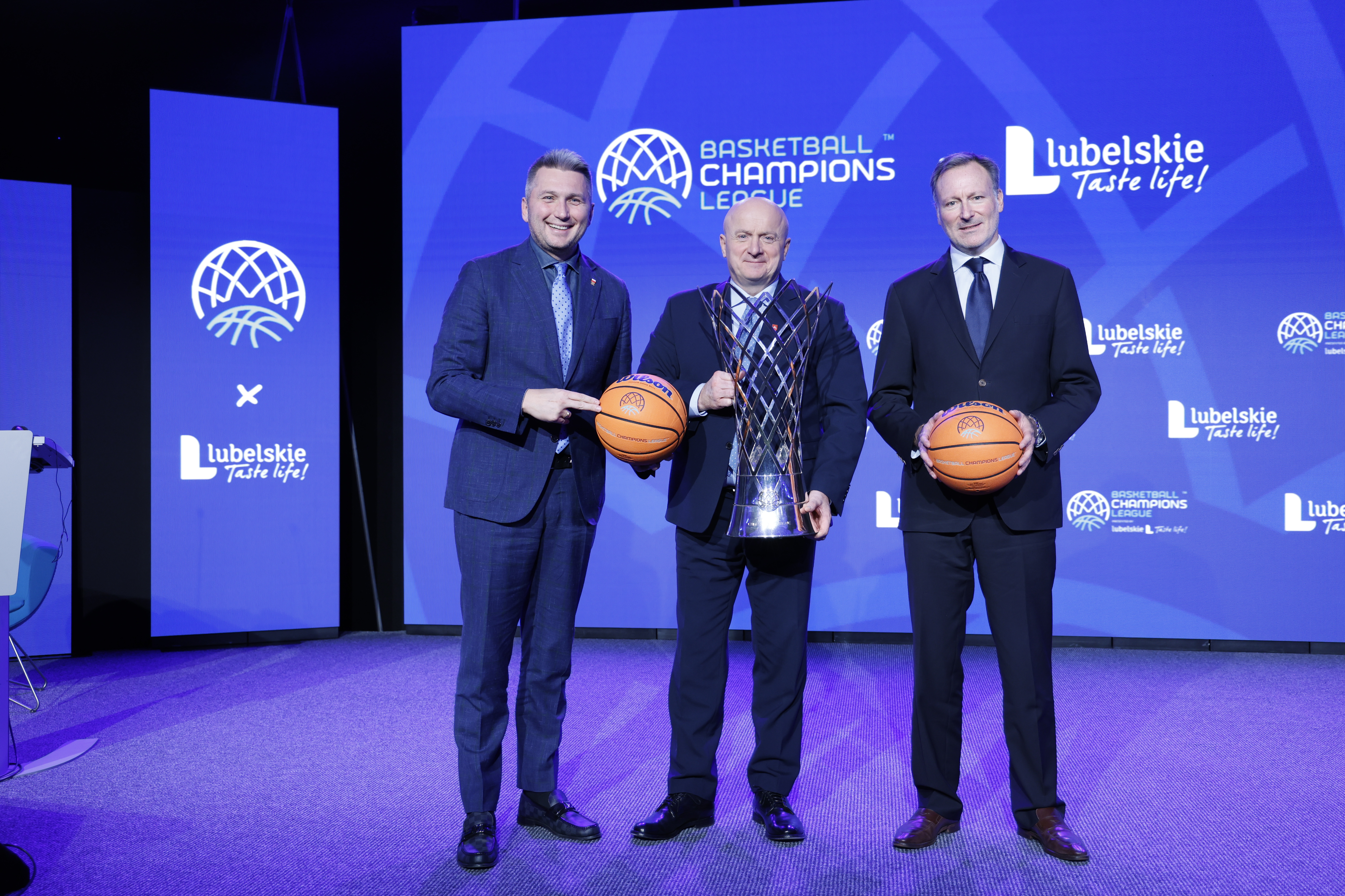 Marszałek w towarzystwie dwóch innych meżczyzn stoi na scenie. Marszałek trzyma w rękach puchar a pozostali panowie trzymają piłki do koszykówki