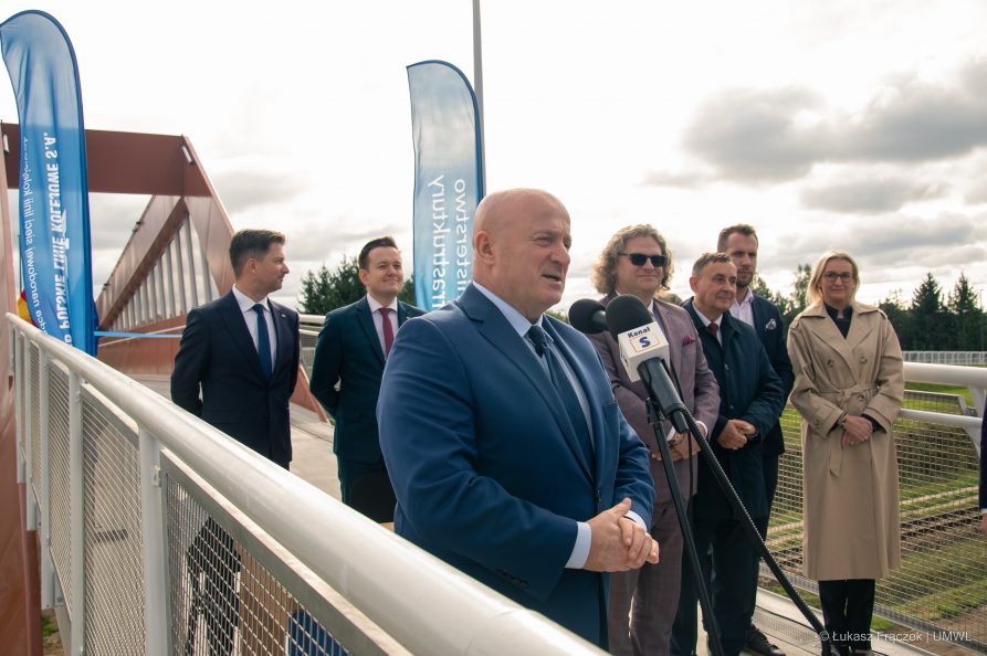 Marszałek Jarosław Stawiarski stoi na kładce w towarzystwie kilku osób i udziela wypowiedzi do mikrofonu