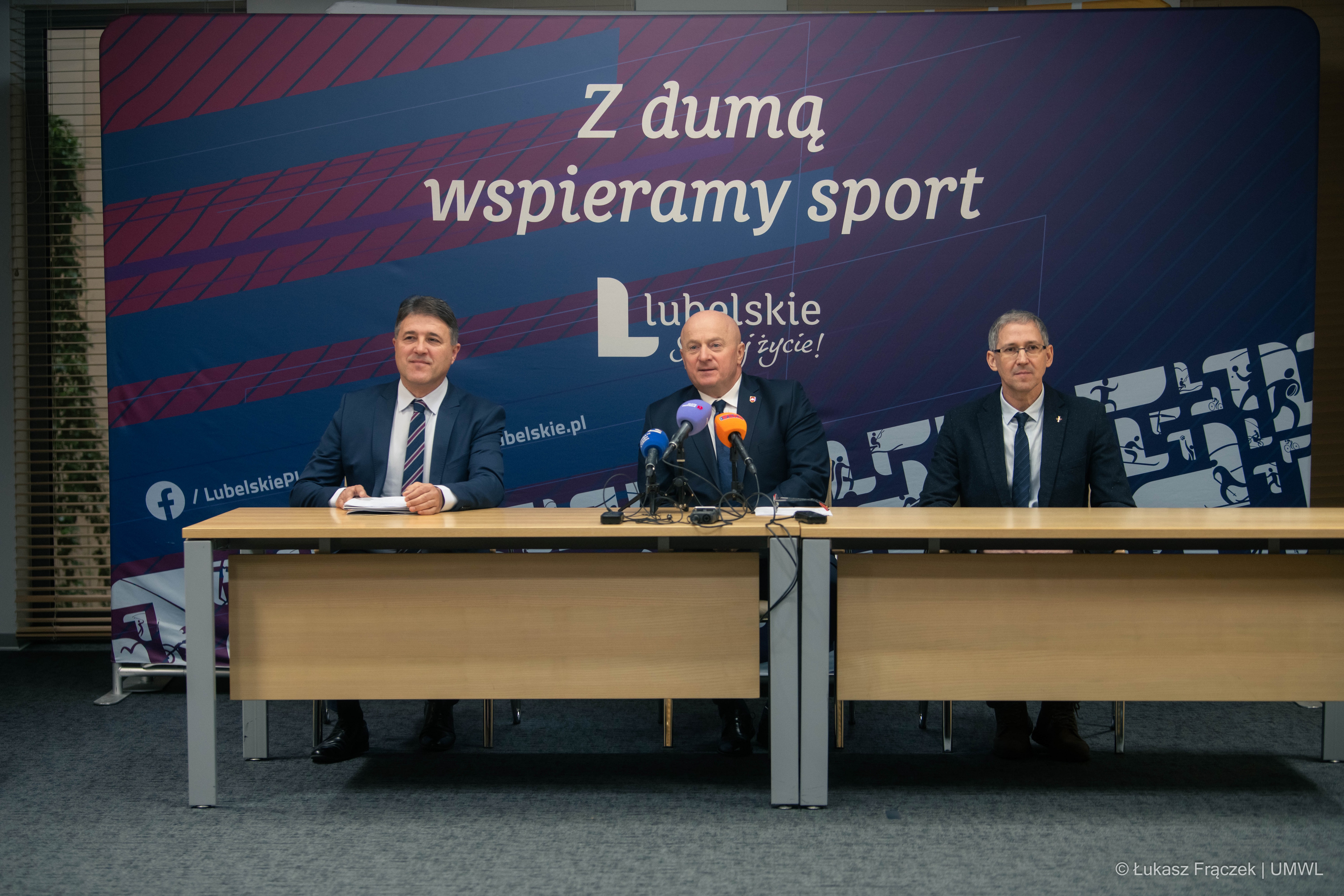 Trzech mężczyzn siedzi przy stoliku, wśród nich obecny jest Marszałek Województwa Lubelskiego. Za nimi znajduje się ścianka z napisem Z dumą wspieramy sport