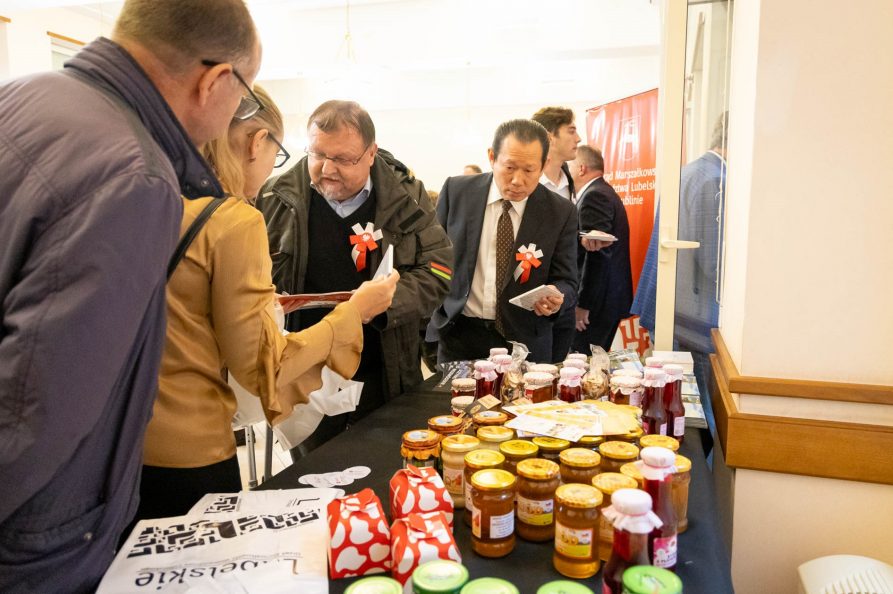 Grupa ludzi stojących wokół stołu ze słoikami dżemu, miodu i soków.