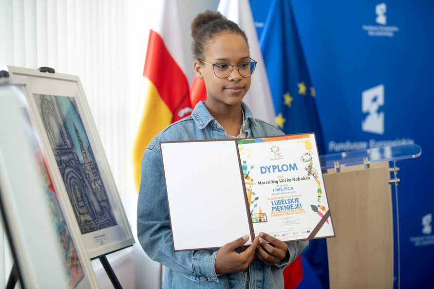 Dziewczyna z dyplomem obok własnej pracy, na tle flag