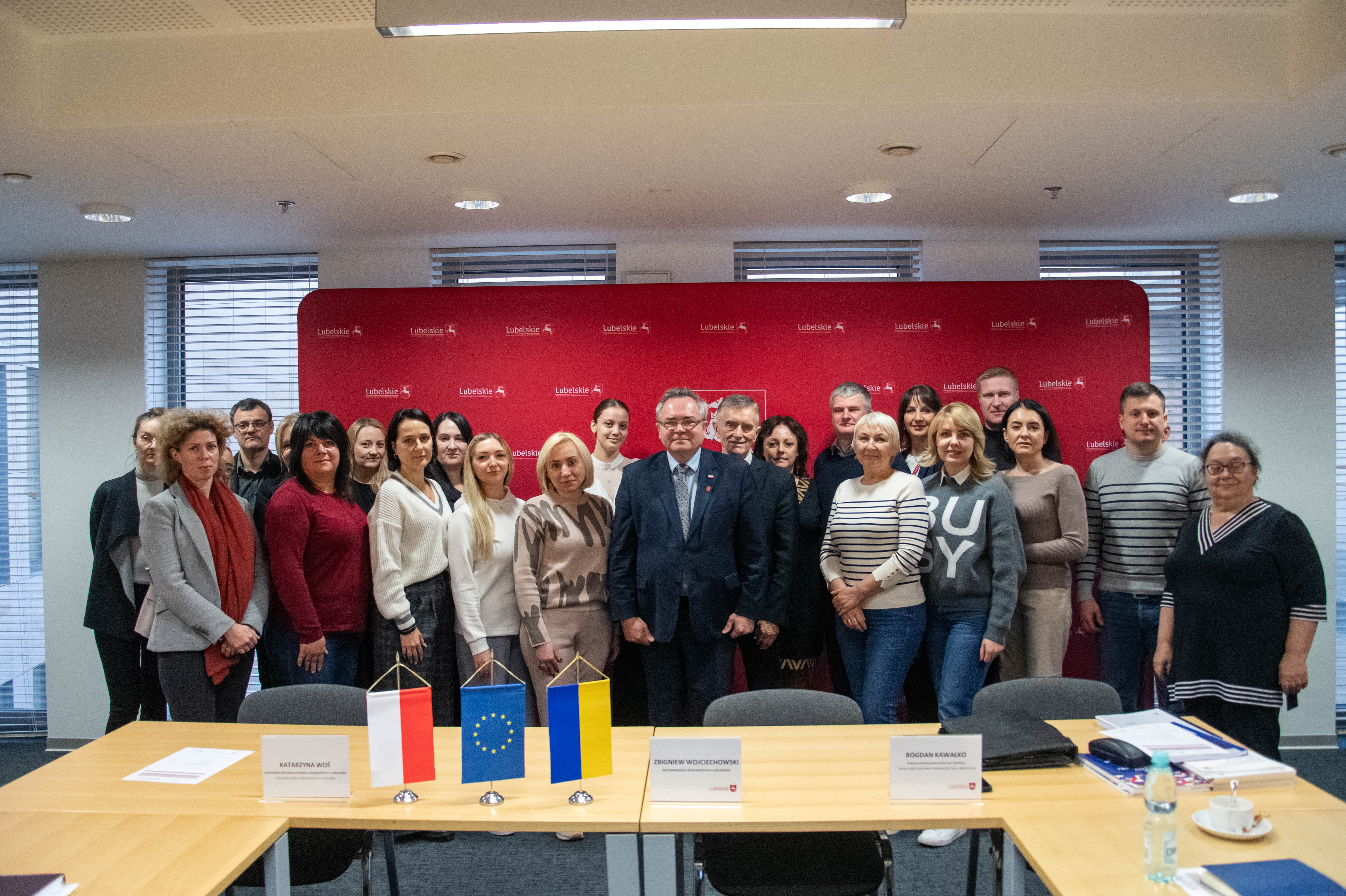 Wciemarszałek Zbigniew Wojciechowski pozuje do zdjęcia wraz z uczestnikami spotkania , łacznie na zdjęciu znajduje się kilkadziesiąt osób stojacych w sali konferencyjnej na tle ścianki