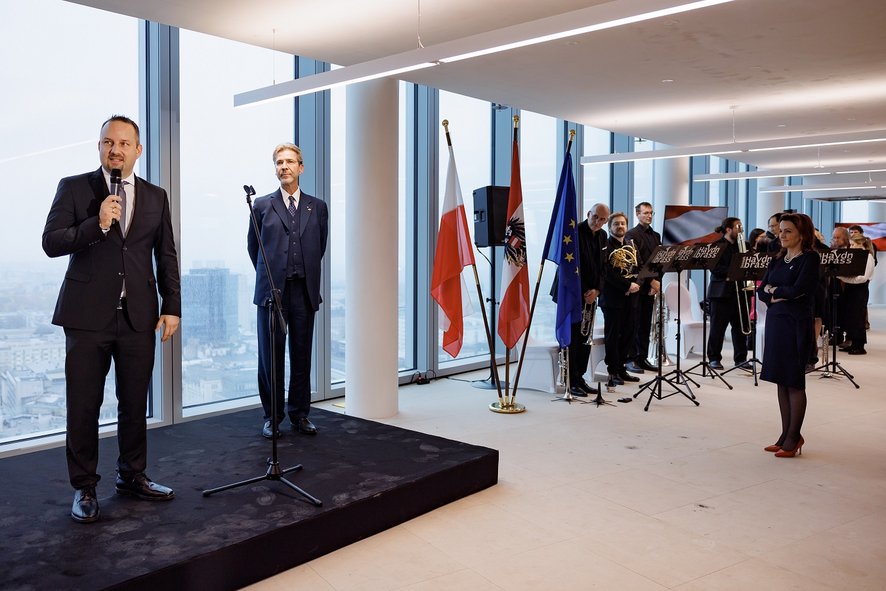 Widok na kilka osób stojących w pomeiszczeniu na wysokim piętrze wieżowca. Dwóch mężczyzn stoi na ustawionym podejście obok nich znajduje się stojak z ustawionymi flagami polską i austriacką a dalej stoi kilka osób