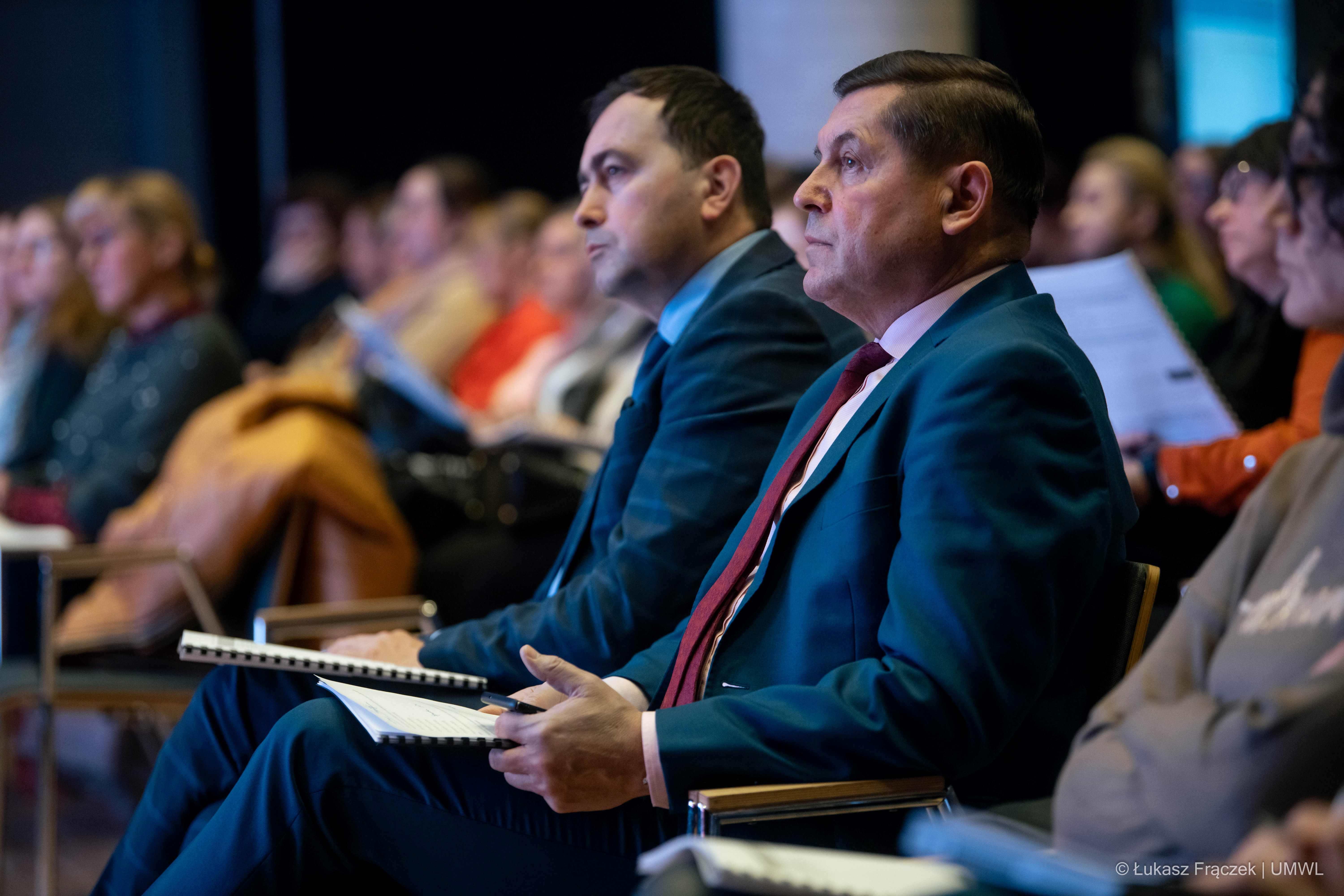 Widok na salę konferencyjną. Na pierwszym planie dwóch słuchaczy konferencji